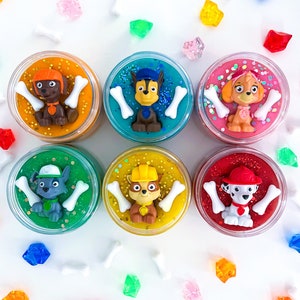 Puede incluir: Seis recipientes circulares llenos de slime de colores, cada uno coronado con una figura de personaje de Paw Patrol y dos dijes blancos en forma de hueso. Los colores del slime incluyen naranja, turquesa, rosa, verde, amarillo y rojo.