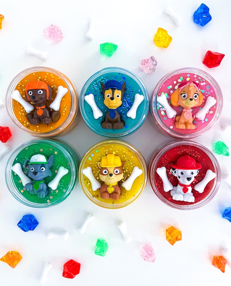 Puede incluir: Seis recipientes circulares llenos de slime de colores, cada uno coronado con una figura de personaje de Paw Patrol y dos dijes blancos en forma de hueso. Los colores del slime incluyen naranja, turquesa, rosa, verde, amarillo y rojo.