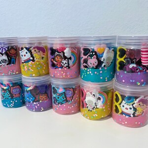 Puede incluir: Una colecci&oacute;n de nueve frascos de pl&aacute;stico transparente llenos de slime de colores y varios dijes de personajes peque&ntilde;os. Cada frasco contiene una combinaci&oacute;n &uacute;nica de colores brillantes, incluyendo amarillo, rosa, azul y morado, mezclados con confeti.