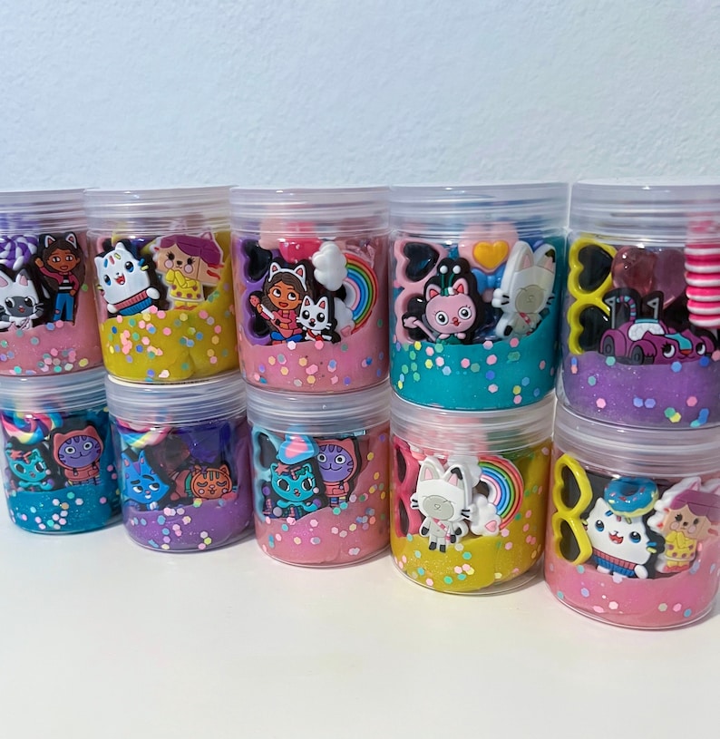 Puede incluir: Una colecci&oacute;n de nueve frascos de pl&aacute;stico transparente llenos de slime de colores y varios dijes de personajes peque&ntilde;os. Cada frasco contiene una combinaci&oacute;n &uacute;nica de colores brillantes, incluyendo amarillo, rosa, azul y morado, mezclados con confeti.