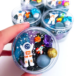 Op de afbeelding: Een doorzichtige plastic pot gevuld met donkerblauwe en zwarte ruimte-thema slijm, voorzien van glitters, een wit astronautfiguurtje, een kleine raket, een kleurrijke planeetbedel, een paars kristal en een gouden glitterbol.