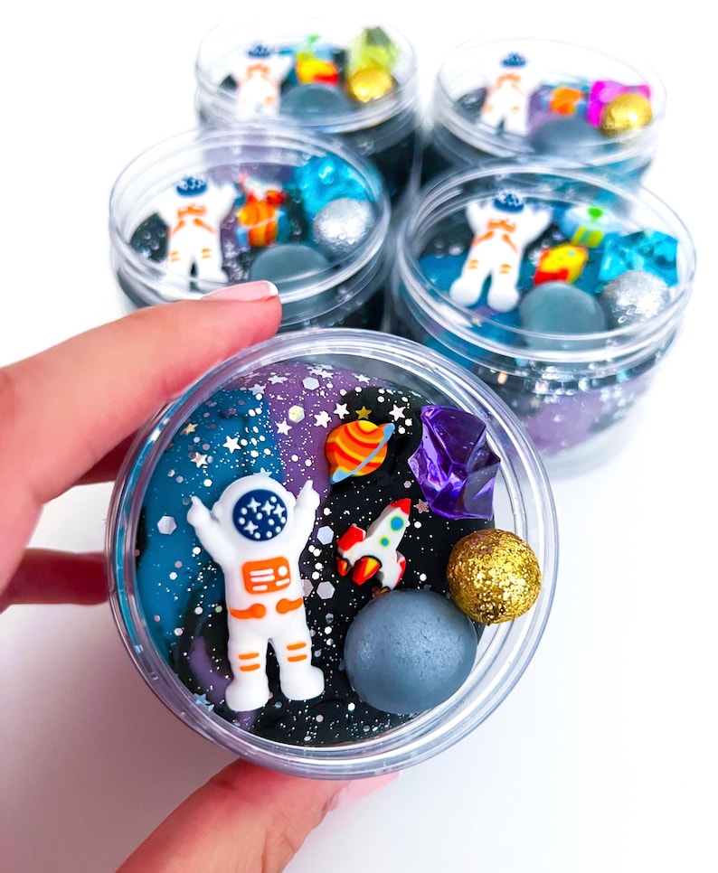 Op de afbeelding: Een doorzichtige plastic pot gevuld met donkerblauwe en zwarte ruimte-thema slijm, voorzien van glitters, een wit astronautfiguurtje, een kleine raket, een kleurrijke planeetbedel, een paars kristal en een gouden glitterbol.