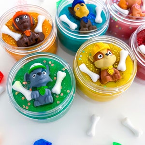 Puede incluir: Cinco frascos de pl&aacute;stico transparente llenos de slime de colores, cada uno coronado con una peque&ntilde;a figura de perro de pl&aacute;stico y dijes blancos en forma de hueso. Los slimes son naranja, turquesa, rosa, verde y amarillo, con purpurina y lentejuelas.