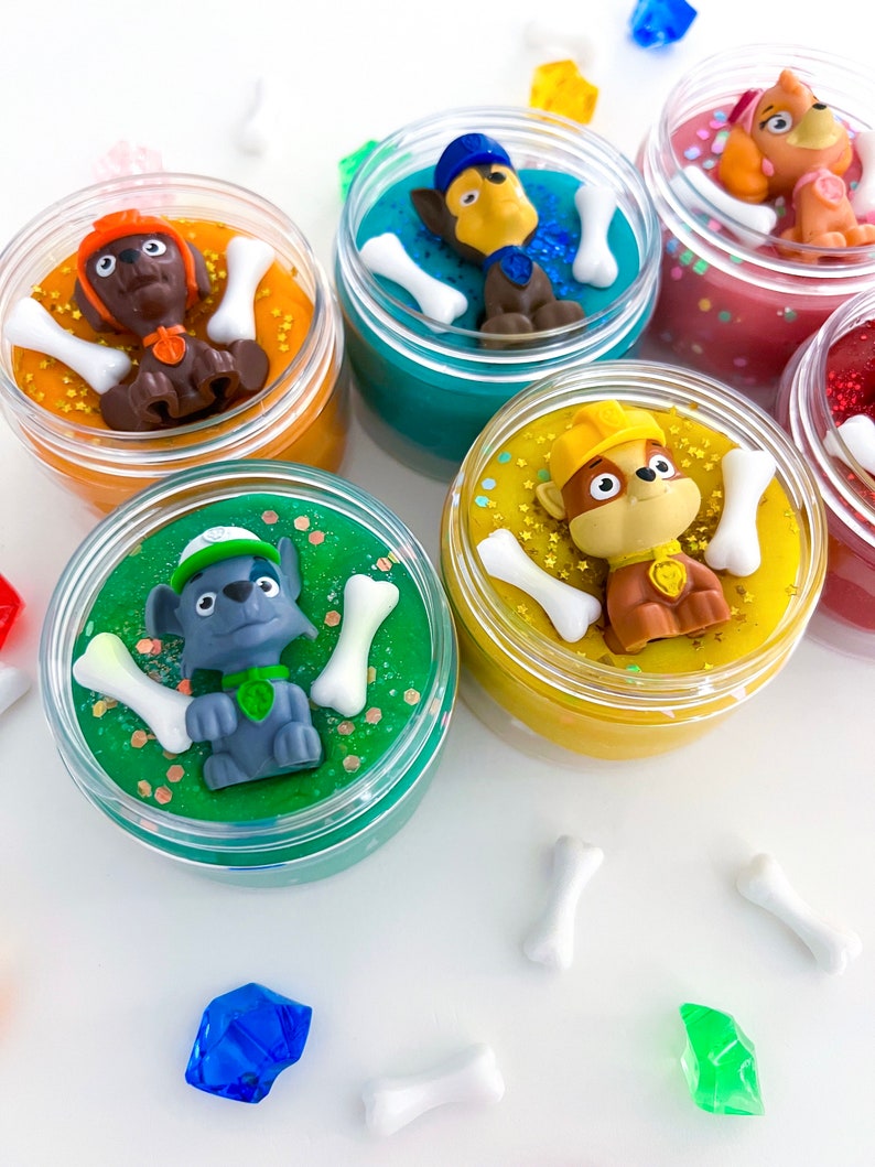 Puede incluir: Cinco frascos de pl&aacute;stico transparente llenos de slime de colores, cada uno coronado con una peque&ntilde;a figura de perro de pl&aacute;stico y dijes blancos en forma de hueso. Los slimes son naranja, turquesa, rosa, verde y amarillo, con purpurina y lentejuelas.