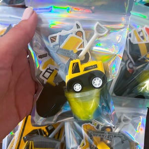 Può includere: Una bustina olografica trasparente richiudibile contenente un camion da cantiere giocattolo giallo e nero con la scritta TRUCK stampata sul lato, una piccola pala in plastica bianca, un contenitore di slime giallo e vari adesivi a tema cantiere.