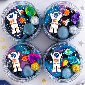 Könnte beinhalten: Vier durchsichtige, runde Behälter gefüllt mit dunkelblauem, schwarzem und violettem Slime. Jeder Behälter enthält eine weiße Astronautenfigur, kleine bunte Planetenanhänger, glitzernde Kugeln und durchscheinende, kristallartige Teile.