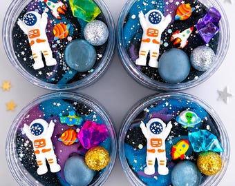 May include: Four clear circular containers filled with dark blue, black, and purple swirled slime. Each container features a white astronaut figurine, small colorful planet charms, glittery spheres, and translucent geometric crystal-like pieces.