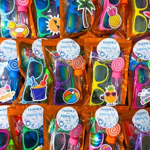 Könnte beinhalten: Eine Sammlung von Partygeschenktüten mit Sonnenbrillen, Seifenblasenflaschen und Lollis. Jede Tüte hat einen Aufkleber mit der Aufschrift "THANKS for popping by!" und sommerliche Aufkleber wie Ananas, Palme und Eimer.