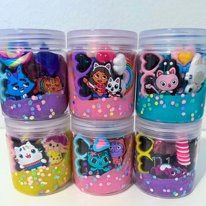 Puede incluir: Seis frascos de pl&aacute;stico transparente llenos de slime de colores y varios dijes con tem&aacute;tica de personajes. Los colores del slime incluyen morado, rosa, azul y amarillo, todos con purpurina iridiscente. Cada frasco presenta dijes de pl&aacute;stico &uacute;nicos como gatos, corazones y arco&iacute;ris.
