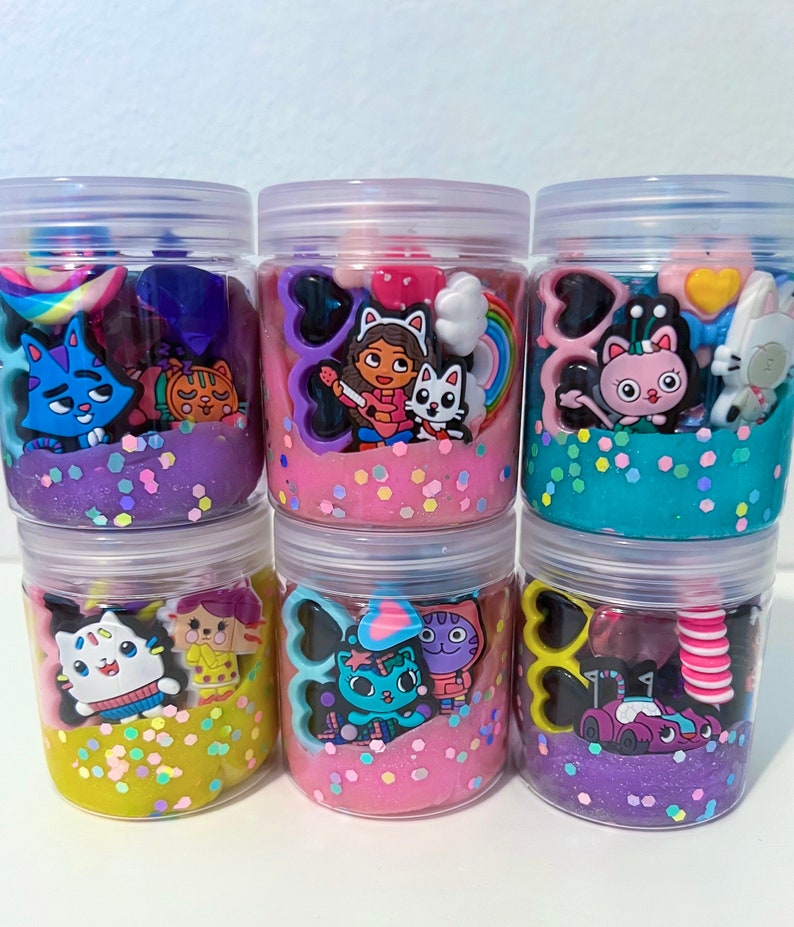 Puede incluir: Seis frascos de pl&aacute;stico transparente llenos de slime de colores y varios dijes con tem&aacute;tica de personajes. Los colores del slime incluyen morado, rosa, azul y amarillo, todos con purpurina iridiscente. Cada frasco presenta dijes de pl&aacute;stico &uacute;nicos como gatos, corazones y arco&iacute;ris.