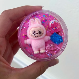 Peut inclure: Un récipient en plastique transparent rempli de slime rose, de paillettes et d'éléments décoratifs. Une figurine de lapin rose est centrée, avec un petit gâteau et des pièces en forme de gemme. Le récipient est tenu par une main.