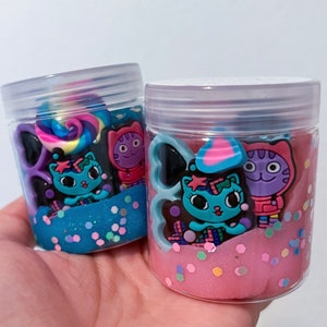 Puede incluir: Dos frascos de pl&aacute;stico transparente llenos de slime de colores. Un frasco contiene slime azul con purpurina plateada y el otro slime rosa con purpurina hexagonal multicolor. Ambos frascos presentan dijes decorativos de gatos de dibujos.