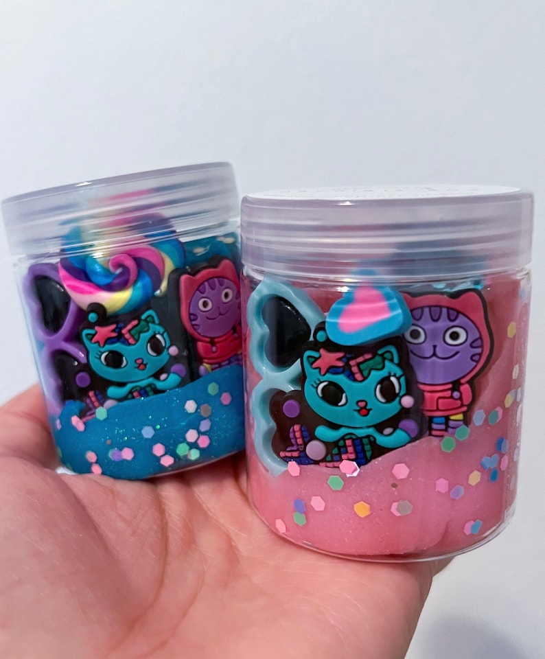 Puede incluir: Dos frascos de pl&aacute;stico transparente llenos de slime de colores. Un frasco contiene slime azul con purpurina plateada y el otro slime rosa con purpurina hexagonal multicolor. Ambos frascos presentan dijes decorativos de gatos de dibujos.