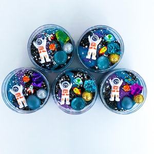 Op de afbeelding: Vijf ronde bakjes gevuld met donkerblauwe en paarse ruimte-thema slijm. Elk bakje bevat een wit astronautfiguurtje, kleine kleurrijke raketbedeltjes, doorschijnende kristalachtige stukjes en glinsterende bollen in goud en zilver.