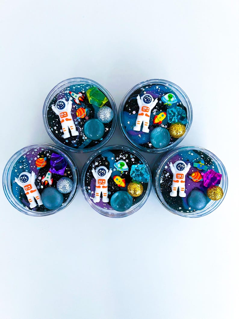 Op de afbeelding: Vijf ronde bakjes gevuld met donkerblauwe en paarse ruimte-thema slijm. Elk bakje bevat een wit astronautfiguurtje, kleine kleurrijke raketbedeltjes, doorschijnende kristalachtige stukjes en glinsterende bollen in goud en zilver.