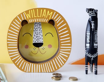 May include: A yellow and brown wooden lion piggy bank with a slot for coins. The lion's face is painted with a smiling expression and the name "William" is written above the slot. The piggy bank is sitting on a white surface with a few coins scattered around it.