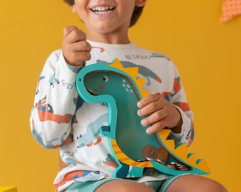 May include: A young child sits on a chair holding a blue and yellow dinosaur-shaped money box. The money box has a slot in the top for coins. The child is smiling and looking up.