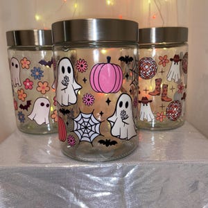 May include: Three glass jars with silver lids, decorated with Halloween-themed decals. The designs include ghosts, flowers, bats, a pink pumpkin, a spider web, disco balls, and cowboy boots. The jars are on a silver surface.