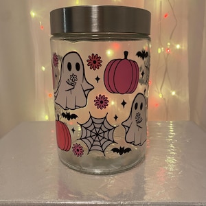 Może przedstawiać: Przezroczysty szklany słoik ze srebrną pokrywką, ozdobiony grafiką o tematyce Halloween. Projekt zawiera r&oacute;żowe dynie, białe duszki trzymające kwiaty, czarne nietoperze i pajęczynę. Słoik stoi na srebrnej powierzchni.