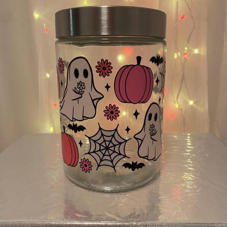 Może przedstawiać: Przezroczysty szklany słoik ze srebrną pokrywką, ozdobiony grafiką o tematyce Halloween. Projekt zawiera r&oacute;żowe dynie, białe duszki trzymające kwiaty, czarne nietoperze i pajęczynę. Słoik stoi na srebrnej powierzchni.