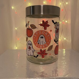 Może przedstawiać: Przezroczysty szklany słoik ze srebrną pokrywką, ozdobiony grafiką o tematyce Halloween. Słoik posiada centralny wz&oacute;r ze słowami "Spooky Vibes" wok&oacute;ł ilustracji ducha. Inne dekoracje to dynie, nietoperze i jesienne liście.