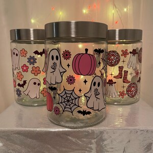 Może przedstawiać: Trzy szklane słoiki ze srebrnymi pokrywkami, ozdobione ilustracjami o tematyce Halloween. Słoiki przedstawiają obrazy duch&oacute;w, dyni, nietoperzy, kwiat&oacute;w i kul dyskotekowych w stylu retro. Słoiki są podświetlane od wewnątrz.