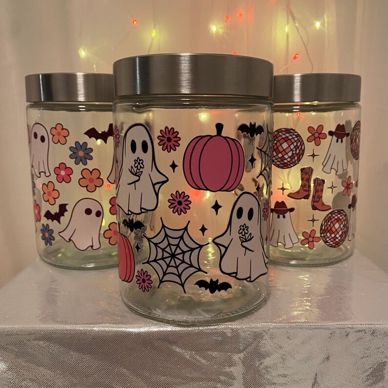 Może przedstawiać: Trzy szklane słoiki ze srebrnymi pokrywkami, ozdobione ilustracjami o tematyce Halloween. Słoiki przedstawiają obrazy duch&oacute;w, dyni, nietoperzy, kwiat&oacute;w i kul dyskotekowych w stylu retro. Słoiki są podświetlane od wewnątrz.
