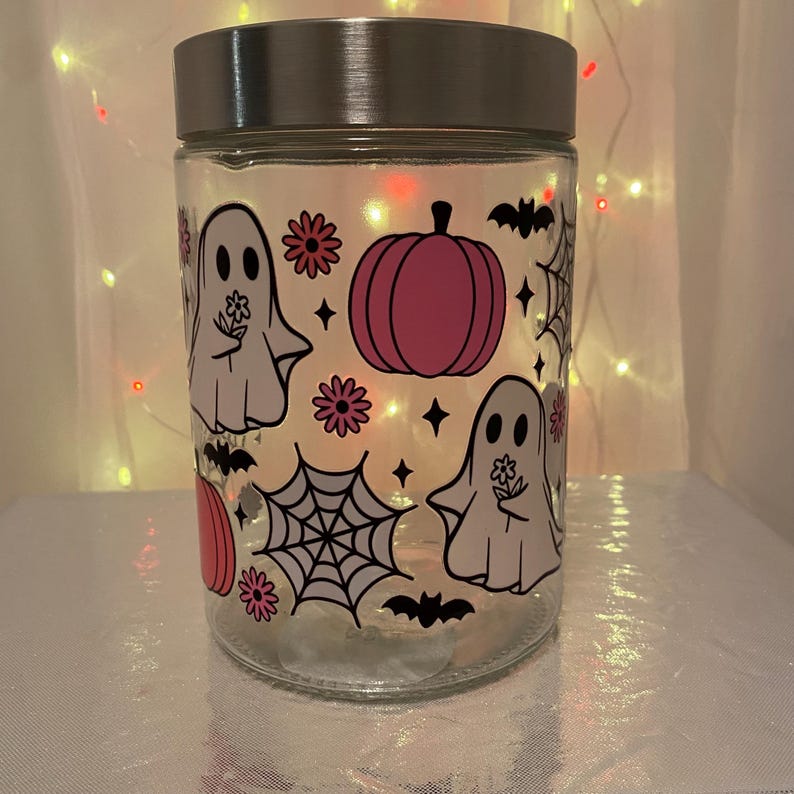 Może przedstawiać: Przezroczysty szklany słoik ze srebrną pokrywką, ozdobiony grafiką o tematyce Halloween. Projekt zawiera r&oacute;żowe dynie, białe duszki trzymające kwiaty, czarne nietoperze, pajęczyny i akcenty gwiazd. Słoik jest podświetlany od wewnątrz.