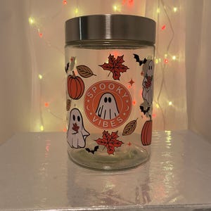 Może przedstawiać: Przezroczysty szklany słoik ze srebrną pokrywką, ozdobiony grafiką o tematyce Halloween. Słoik przedstawia pomarańczowe dynie, czerwone liście klonu, nietoperze i kresk&oacute;wkowe duszki. Na słoiku wydrukowano napis "Spooky Vibes".