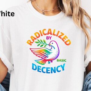 May include: White t-shirt with the text "RADICALIZED BY BASIC DECENCY" in a rainbow arc above a colorful dove holding an olive branch. The dove's wings and tail are in rainbow colors. The word "White" is in the upper left corner.