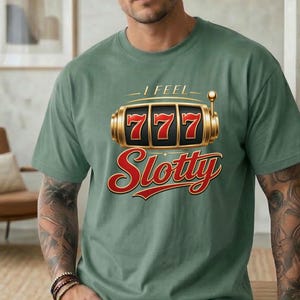 May include: A sage green short-sleeve t-shirt featuring a graphic of a gold slot machine with the number 777 displayed on the reels. Above the graphic is the text I FEEL and below the graphic is the word Slotty in a bold red script font.