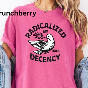 May include: A bright pink t-shirt with the text "RADICALIZED BY BASIC DECENCY" in a circular design. A white dove with olive branch is in the center. The word "Crunchberry" is in the upper left corner.