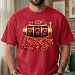 May include: A red short-sleeved t-shirt featuring a graphic of a gold slot machine with the number 777 displayed on the reels. Above the slot machine, the text reads I FEEL, and below it, the word Slotty is written in a large red script font.