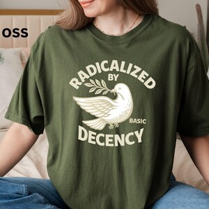 May include: Moss green t-shirt with the text "RADICALIZED BY BASIC DECENCY" and a white dove graphic holding an olive branch. The shirt is a relaxed fit and made of a soft material.