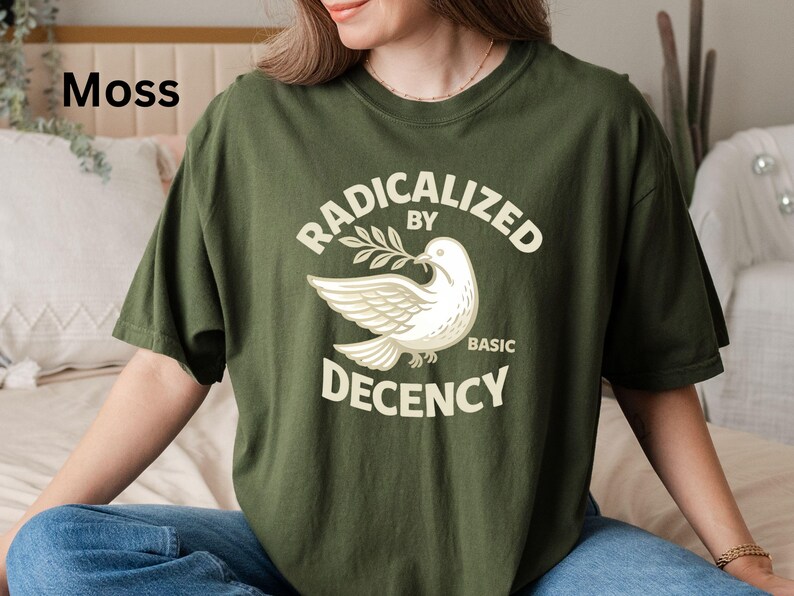 May include: Moss green t-shirt with the text "RADICALIZED BY BASIC DECENCY" and a white dove graphic holding an olive branch. The shirt is a relaxed fit and made of a soft material.