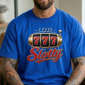 May include: A royal blue short-sleeve t-shirt featuring a graphic of a gold slot machine with three red sevens in the center. Above the slot machine is the text I FEEL in white, and below it is the word Slotty in a large red cursive font.