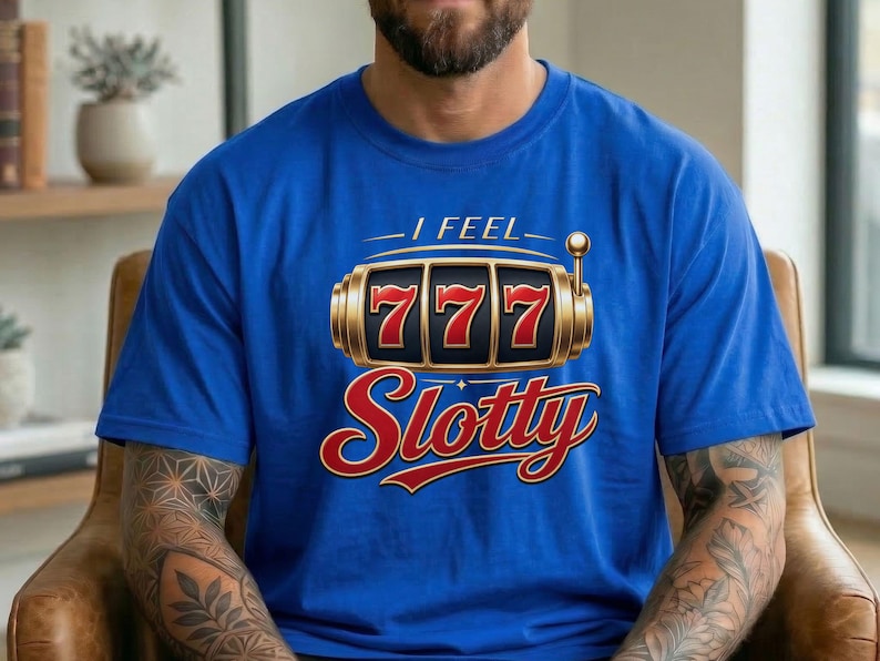 May include: A royal blue short-sleeve t-shirt featuring a graphic of a gold slot machine with three red sevens in the center. Above the slot machine is the text I FEEL in white, and below it is the word Slotty in a large red cursive font.