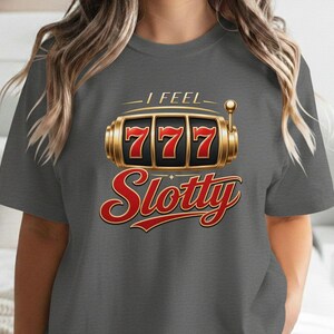 May include: A charcoal gray short-sleeve t-shirt featuring a graphic of a gold slot machine with the number 777 displayed in red. Above the slot machine is the text I FEEL in gold, and below it is the word Slotty written in a large red script font.