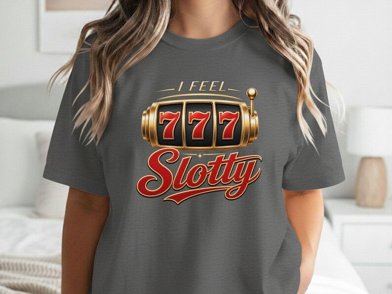 May include: A charcoal gray short-sleeve t-shirt featuring a graphic of a gold slot machine with the number 777 displayed in red. Above the slot machine is the text I FEEL in gold, and below it is the word Slotty written in a large red script font.