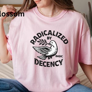 May include: A light pink t-shirt with the text "RADICALIZED BY BASIC DECENCY" in black, featuring a dove holding an olive branch. The word "Blossom" is printed in black in the upper left corner.
