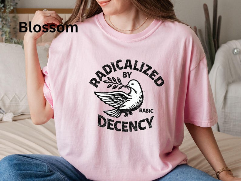 May include: A light pink t-shirt with the text "RADICALIZED BY BASIC DECENCY" in black, featuring a dove holding an olive branch. The word "Blossom" is printed in black in the upper left corner.