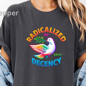 May include: Dark gray t-shirt with the text "RADICALIZED BY BASIC DECENCY" in rainbow colors. A white dove with rainbow wings and a green olive branch is in the center of the design. The shirt has rolled-up sleeves.
