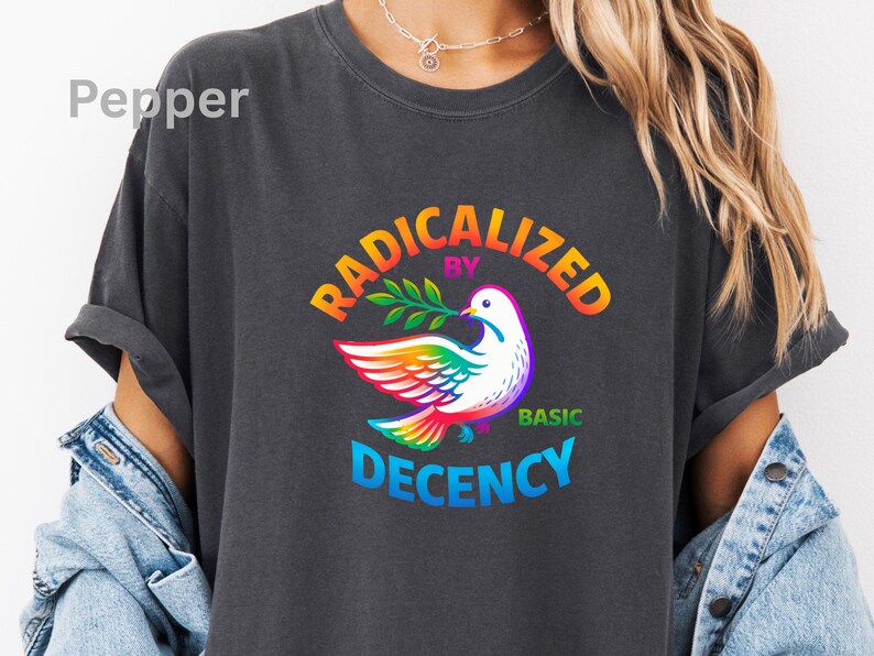 May include: Dark gray t-shirt with the text "RADICALIZED BY BASIC DECENCY" in rainbow colors. A white dove with rainbow wings and a green olive branch is in the center of the design. The shirt has rolled-up sleeves.