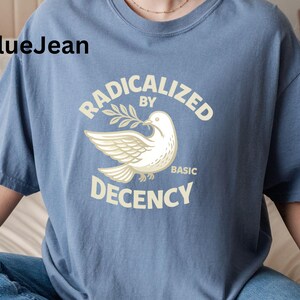 May include: Blue crew neck t-shirt with the text "RADICALIZED BY BASIC DECENCY" and a dove graphic holding an olive branch. The shirt is a muted blue color.
