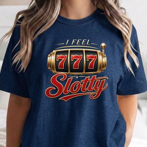 May include: A navy blue short-sleeve t-shirt featuring a graphic of a gold slot machine with three red sevens in the windows. Above the slot machine is the text I FEEL and below it is the word Slotty in a large red cursive font.