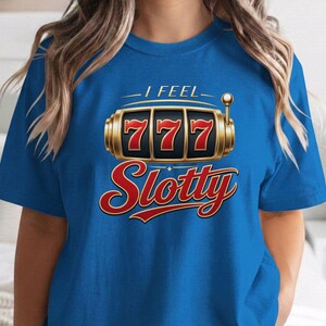 May include: A bright blue short-sleeve t-shirt featuring a graphic of a gold slot machine with three red sevens in the display. Above the slot machine, the text reads I FEEL in white, and below it, the word Slotty is printed in large red script.