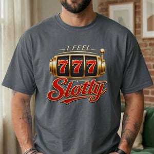 May include: A charcoal gray t-shirt featuring a graphic of a gold slot machine with the number 777 displayed on the reels. Above the slot machine is the text I FEEL and below it is the word Slotty in a bold, red, cursive-style font.