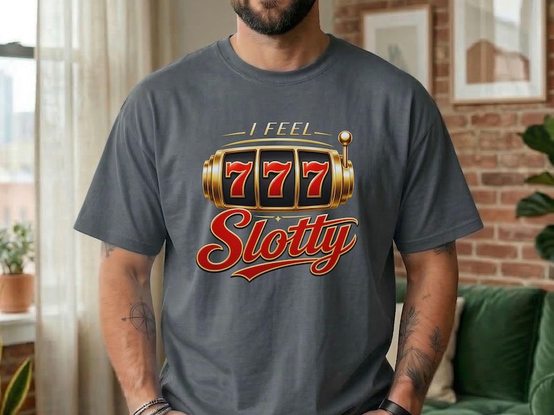 May include: A charcoal gray t-shirt featuring a graphic of a gold slot machine with the number 777 displayed on the reels. Above the slot machine is the text I FEEL and below it is the word Slotty in a bold, red, cursive-style font.