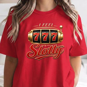May include: A bright red short-sleeved t-shirt featuring a graphic of a gold slot machine with three sevens displayed in the center. Above the slot machine is the text I FEEL and below it is the word Slotty in a bold red and gold font.