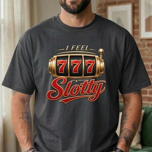 May include: A charcoal gray t-shirt featuring a graphic of a gold slot machine with the number 777 displayed on the reels. Above the slot machine is the text I FEEL and below it is the word Slotty in a large, bold, red cursive font.